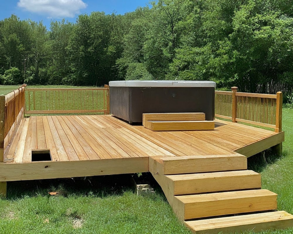A spacious wooden deck featuring a built-in hot tub, surrounded by lush greenery and clear skies